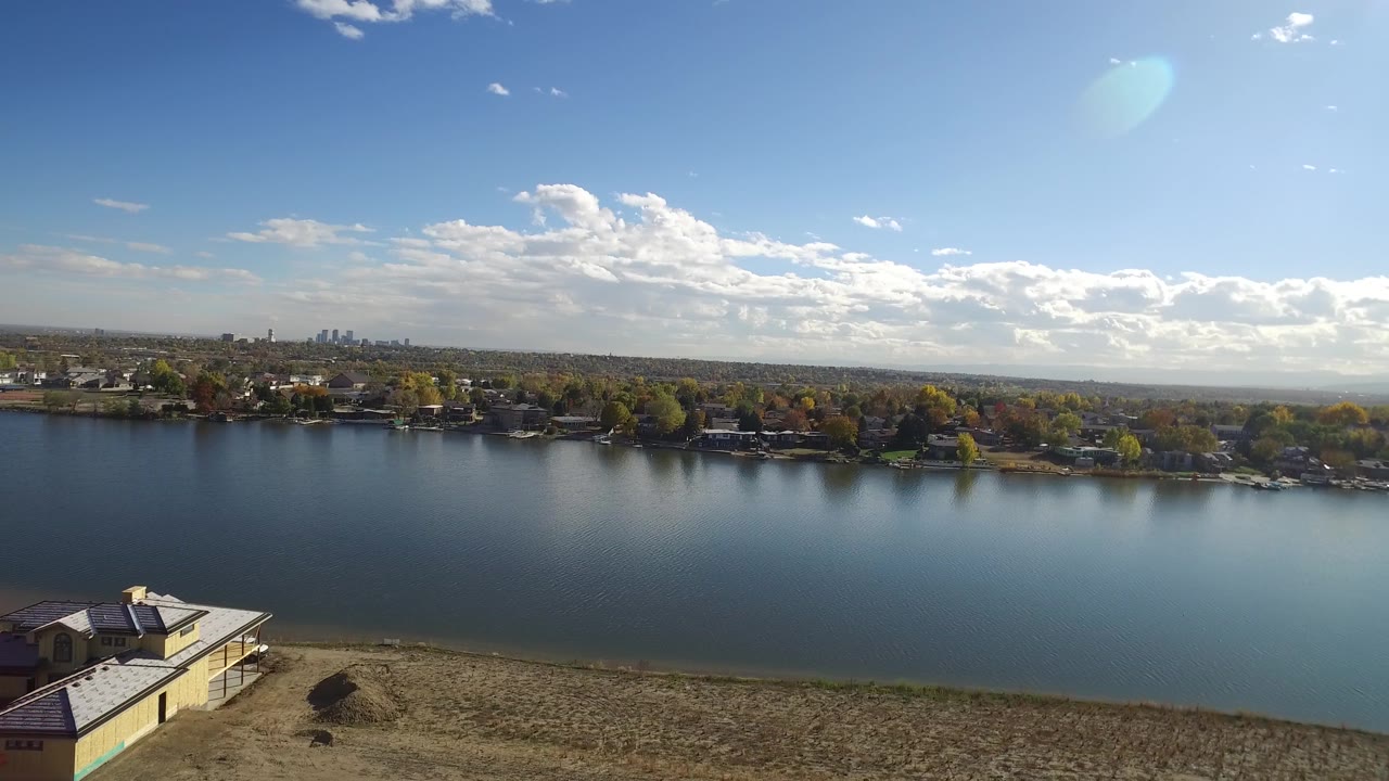 Hidden Lake, Colorado frame with the water centered, the foreground house still partly visible at lower left, far-bank homes and autumn-toned trees on the horizon, scattered clouds, and no visible people.