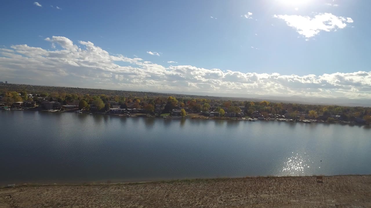 Wide aerial view of a lake with sun glare, shoreline trees, and houses across the water.