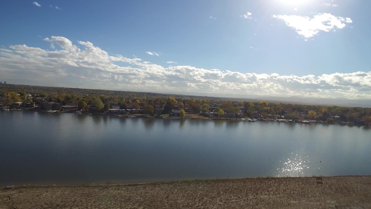Aerial frame of Hidden Lake, Colorado, showing reflective open water, a tree-lined residential shore, scattered clouds, distant hills, and no visible people.