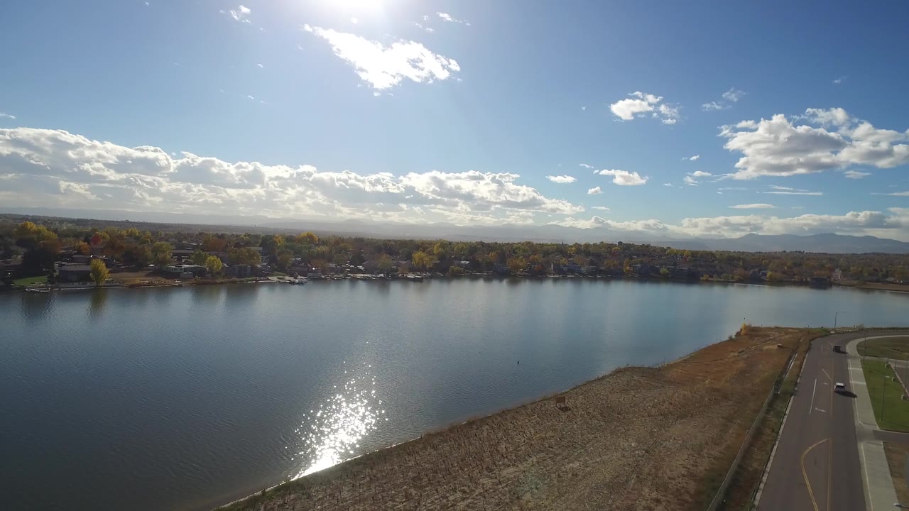 Opening aerial frame over Hidden Lake, Colorado, with bright sun glare on blue water, dry shoreline in front, a road at right, distant trees and mountains, and no visible people.