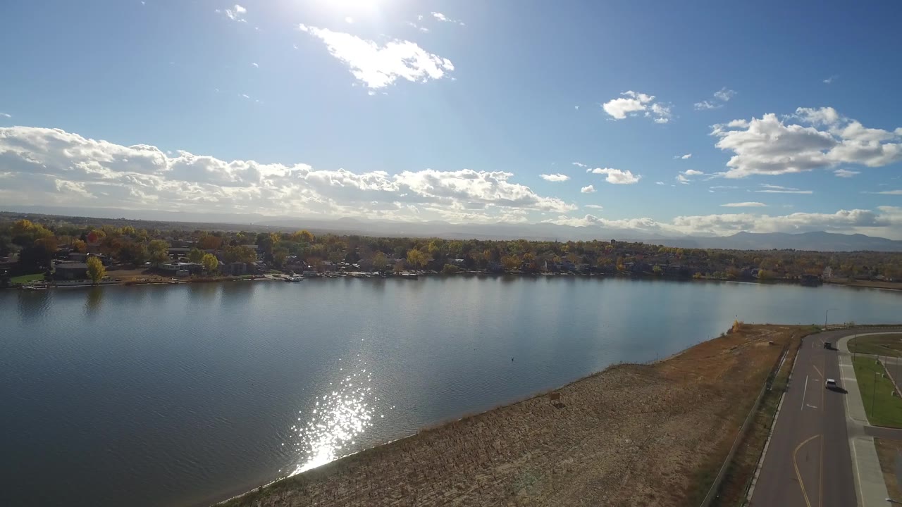 Aerial frame over Hidden Lake, Colorado, with the water reflection lower in frame, dry shoreline crossing the foreground, a road on the right edge, and no visible people.