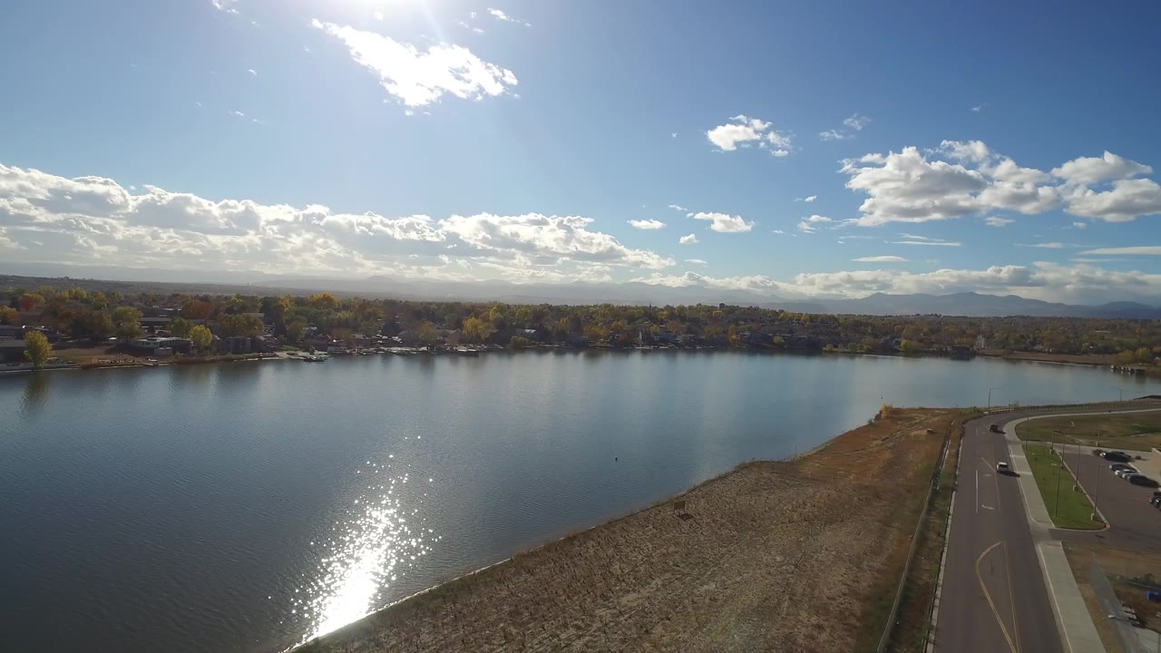 Lake and tree-lined opposite shore under a bright sky, with road and open shoreline at right.