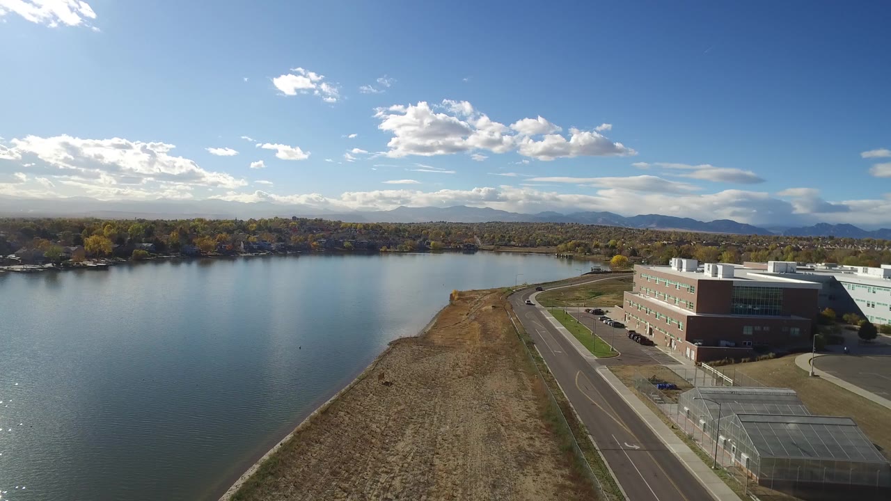 Opening aerial frame over Hidden Lake, Colorado, with calm water on the left, a dry shoreline road below, low buildings to the right, mountains on the horizon, and no visible people.