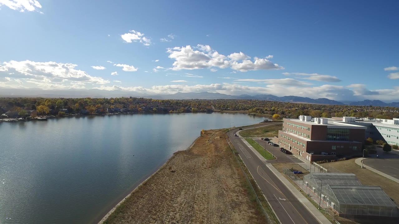 Aerial view at 0:01 over Hidden Lake, Colorado, with the lake filling the left side, the shoreline road leading toward low buildings, scattered clouds overhead, and no visible people.