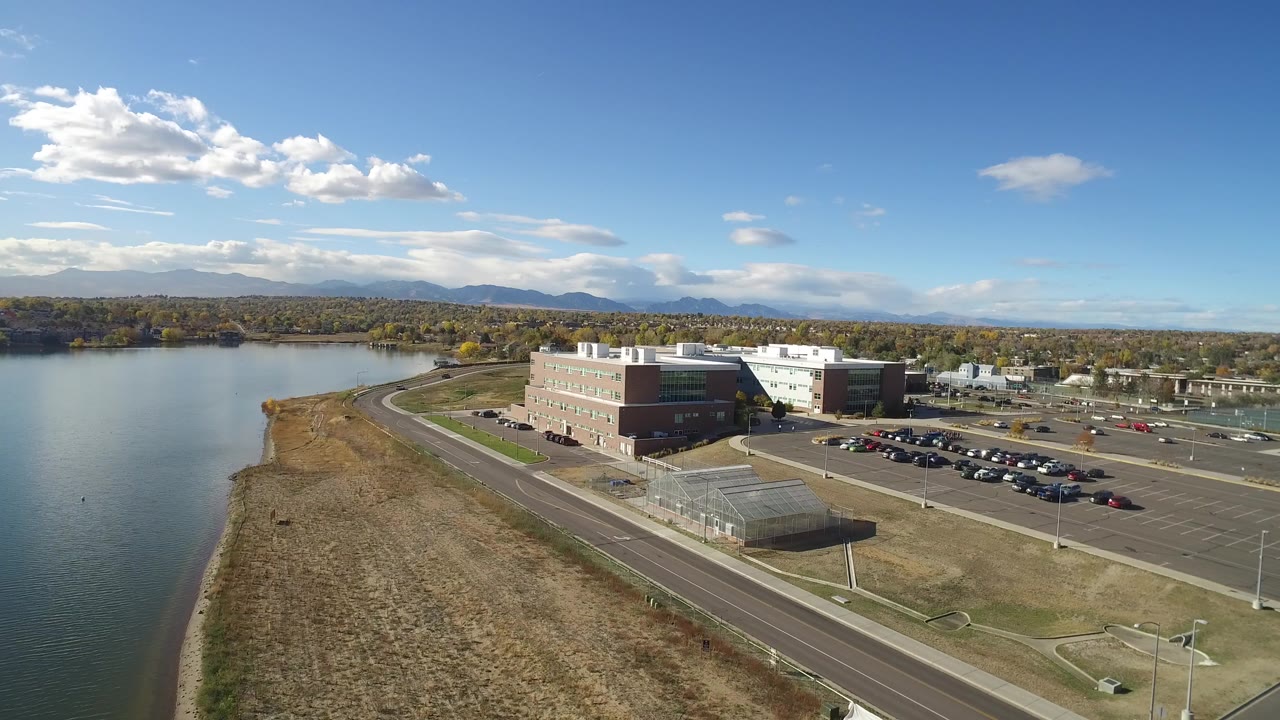 Opening aerial frame over Hidden Lake, Colorado, showing water at left, dry shoreline grass, a modern office building, parking lots, distant mountains, and no visible people.