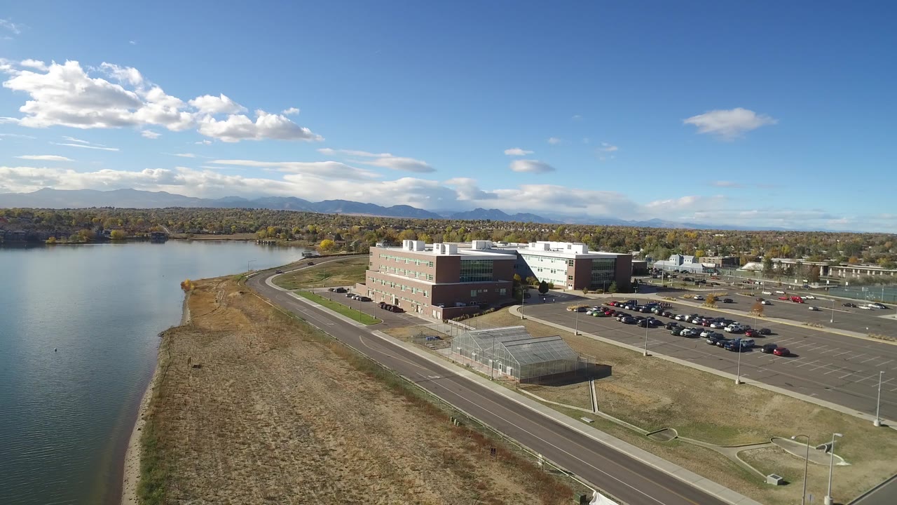 A second wide aerial view in Hidden Lake, Colorado keeps the lake at left and the office building centered, with parking rows, roads, trees, mountains, and no visible people.