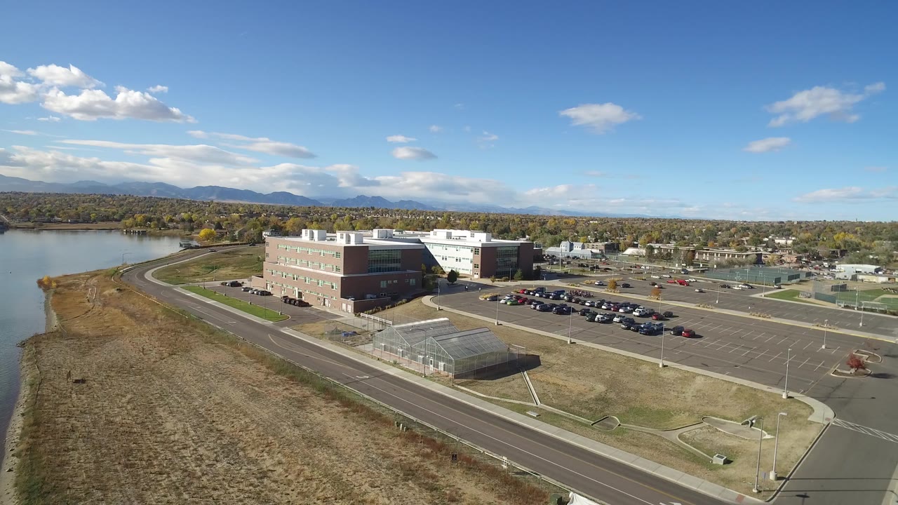 Drone frame over Hidden Lake, Colorado, with the main building centered, parked cars in the lot, a greenhouse-like structure near the road, mountains in the distance, and no visible people.