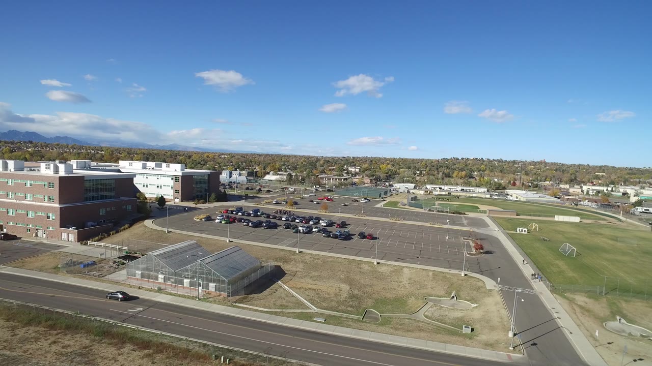 Hidden Lake, Colorado aerial frame with the brick-and-white building at left, a large mostly open parking lot, sports fields to the right, and distant houses along the horizon.