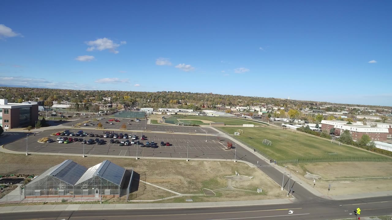 Aerial view of road, greenhouses, parking lot, fields, and low buildings under blue sky.