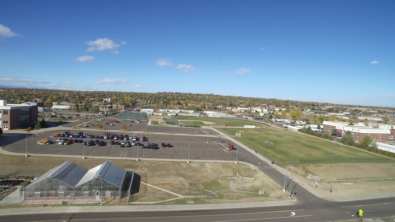 Aerial view in Hidden Lake, Colorado, looking across a foreground road, dry grass, parking rows, sports fields, and low buildings, with no visible people.