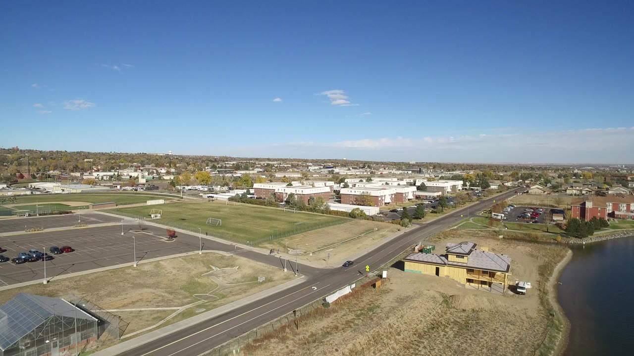 Aerial view of a road, sports fields, parking lots, buildings, and a lake edge under blue sky.
