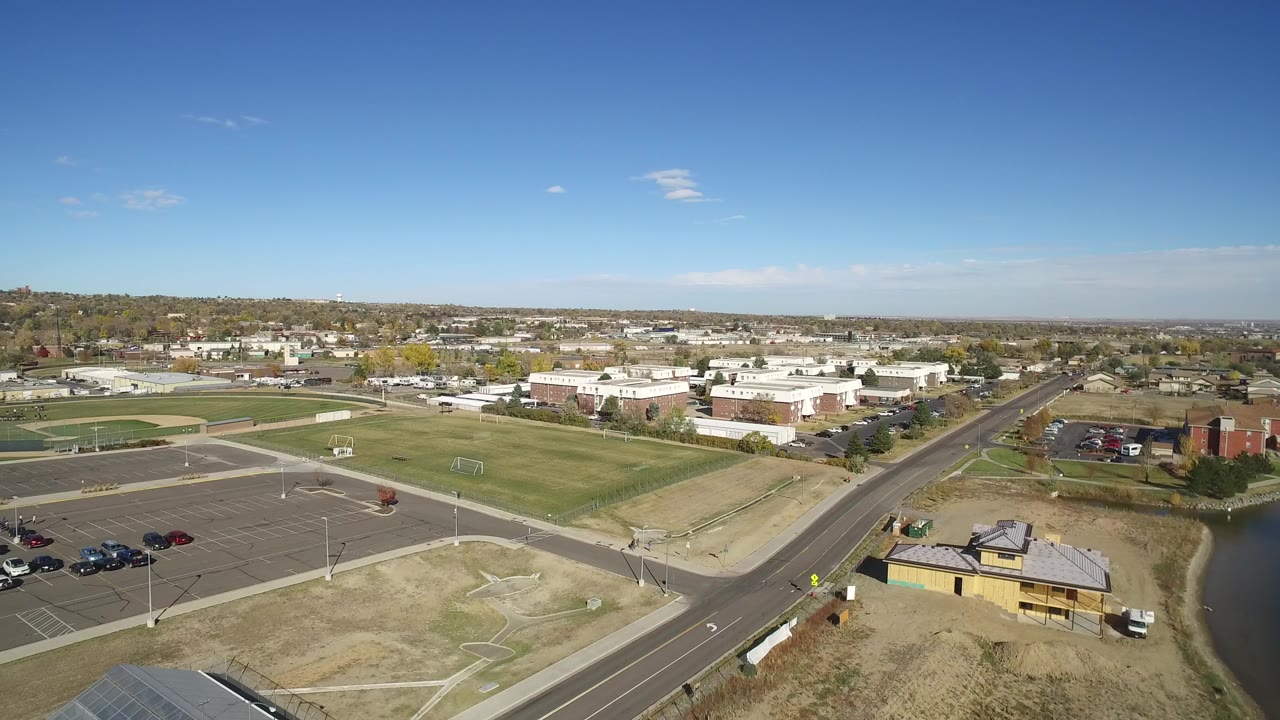 Opening aerial frame over Hidden Lake, Colorado, showing a road intersection, green fields, parking lots, low buildings, the lake at right, and no visible people.