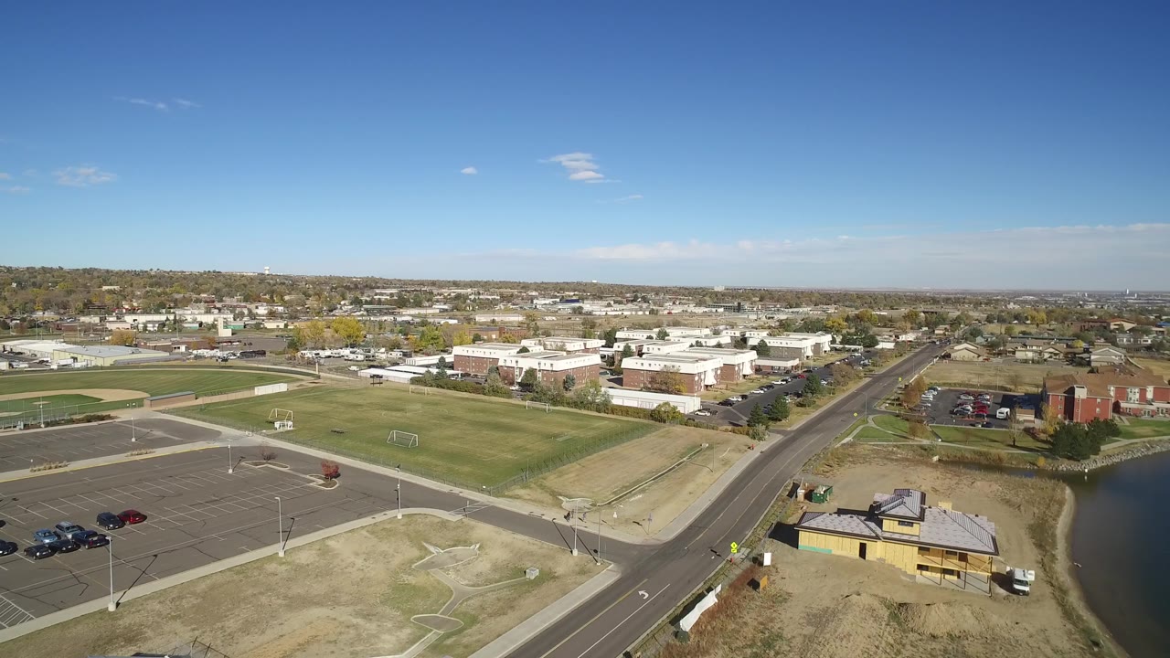 Wide aerial frame with athletic fields, a road corridor, low buildings, and a lake shoreline.