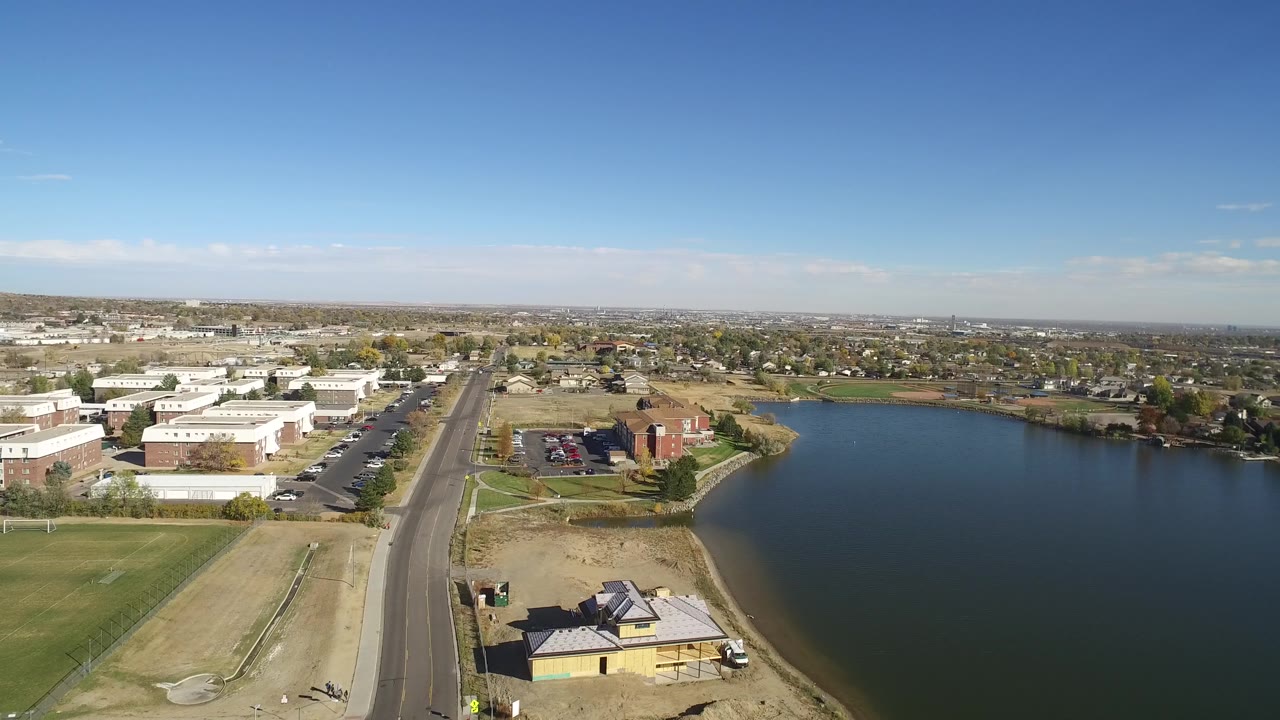 Drone-like view across a lake with road, parking areas, and homes on the shore