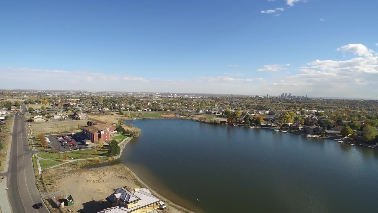 Wide aerial frame from Hidden Lake, Colorado with the lake filling most of the view, sandy shoreline below, brick buildings at left, homes across the far bank, and no visible people.