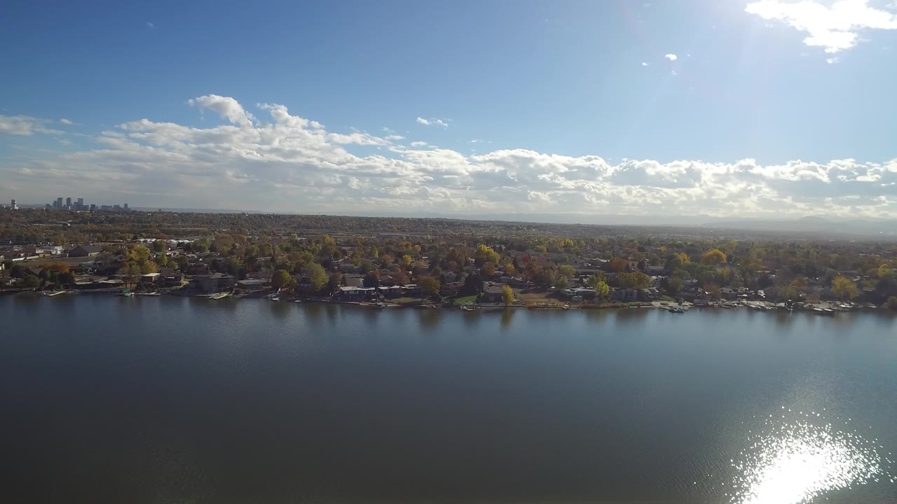 Opening frame of Hidden Lake, Colorado, with dark blue water in the foreground, bright sun glare near the lower edge, shoreline homes and trees across the middle, distant buildings on the horizon, and no visible people.