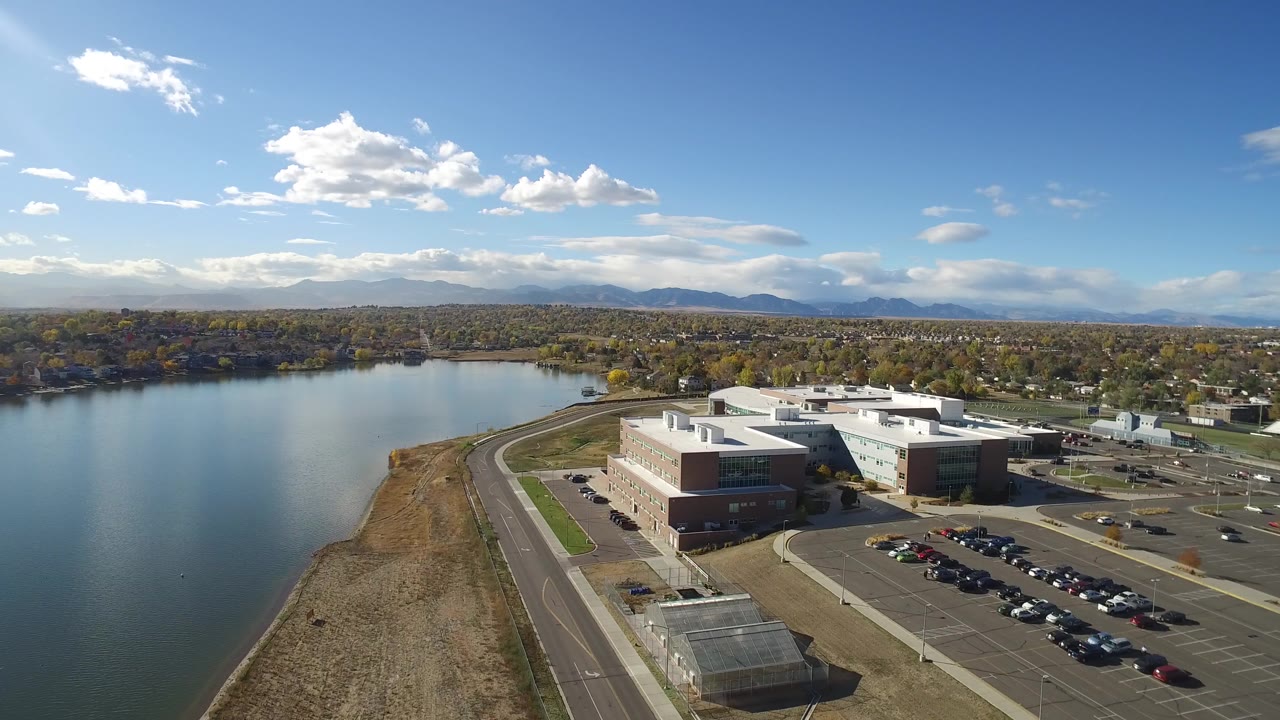 Aerial view of a lake, shoreline road, modern building complex, parking lot, and distant mountains.