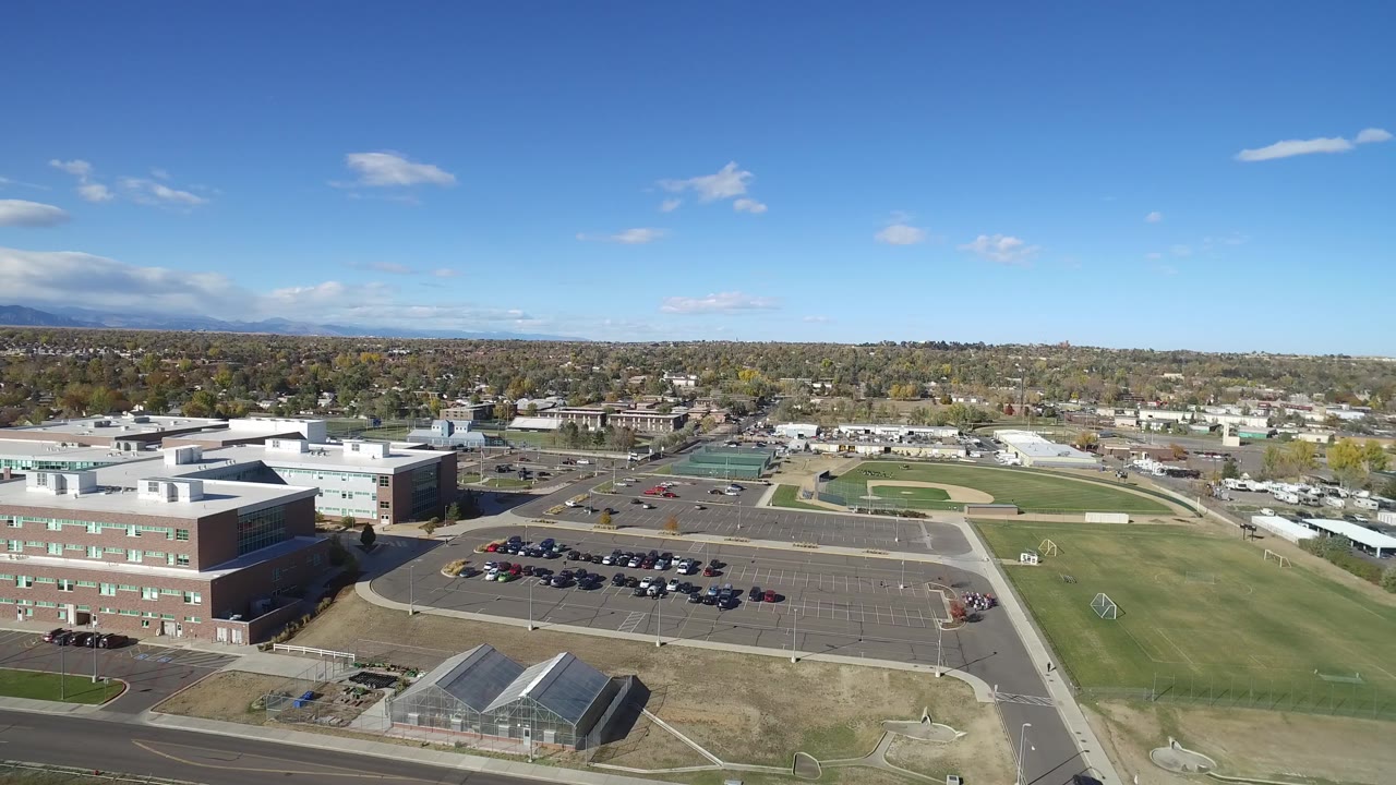Second aerial frame in Hidden Lake, Colorado, with brick buildings at left, parked cars below, baseball fields beyond the lot, tree-lined neighborhoods, and no visible people.