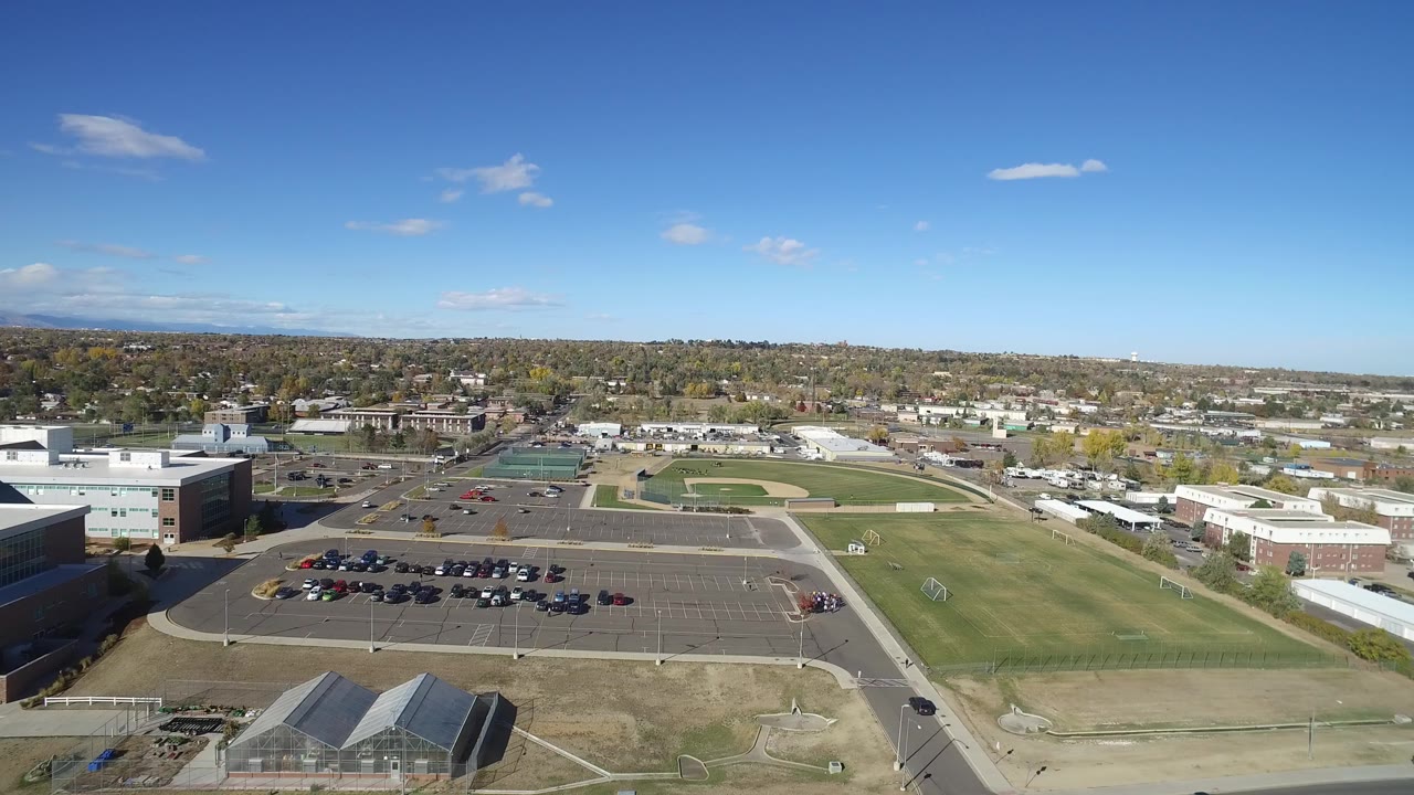 Aerial frame from Hidden Lake, Colorado showing a green field beside a large parking lot, low administrative buildings, distant homes, and no visible people.