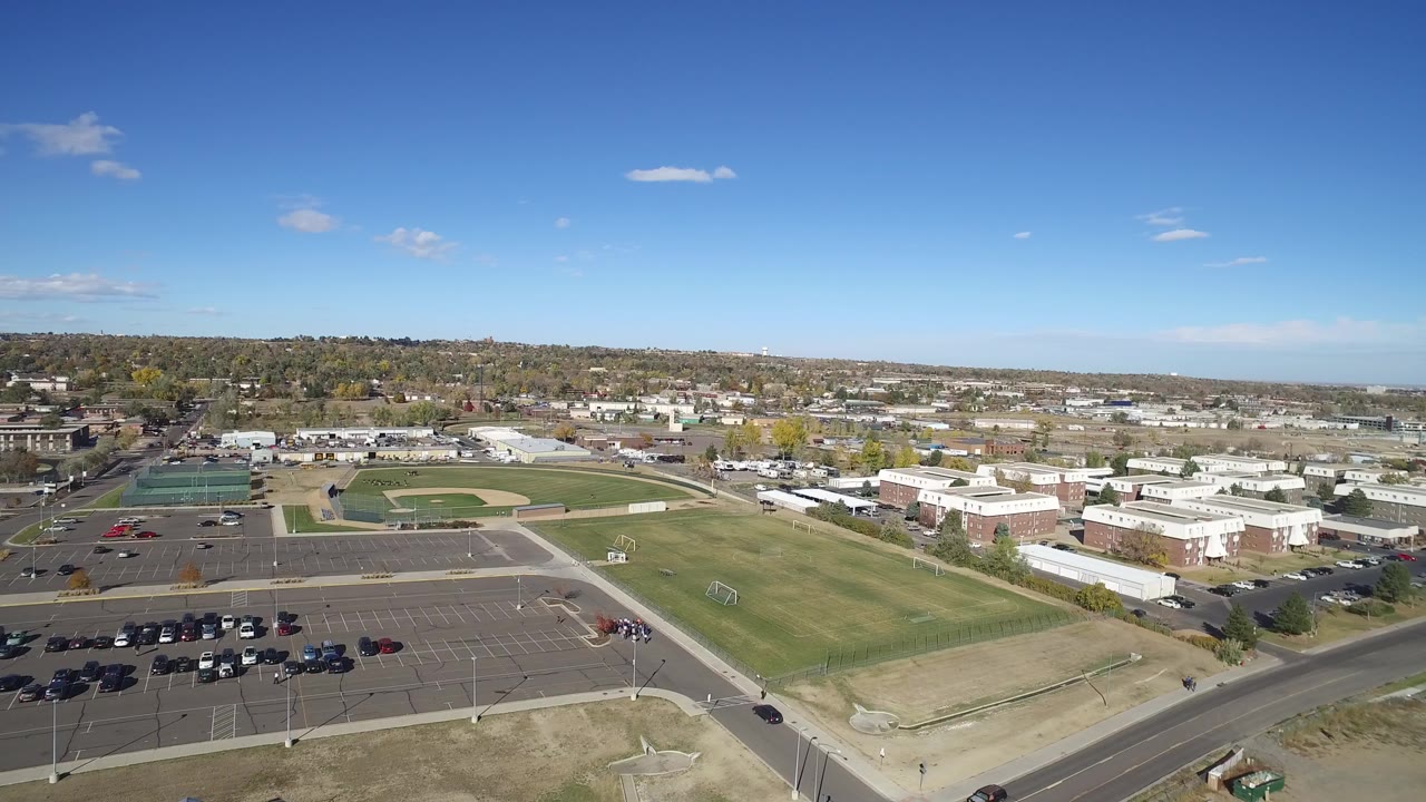 Opening aerial frame over Hidden Lake, Colorado showing a parking lot, green sports fields, low buildings, roads, distant rooftops, and no visible people.
