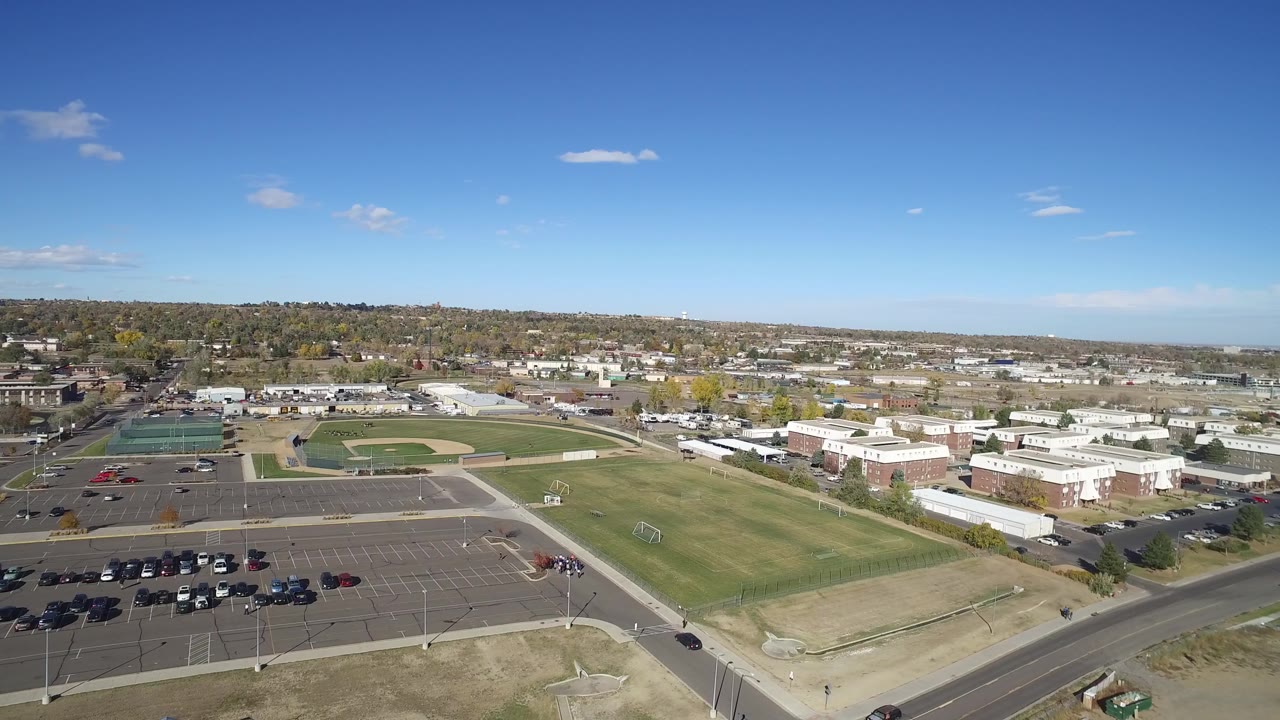 Aerial frame in Hidden Lake, Colorado with the parking lot lower in view, a baseball field to the left, school-like buildings in the midground, and no visible people.