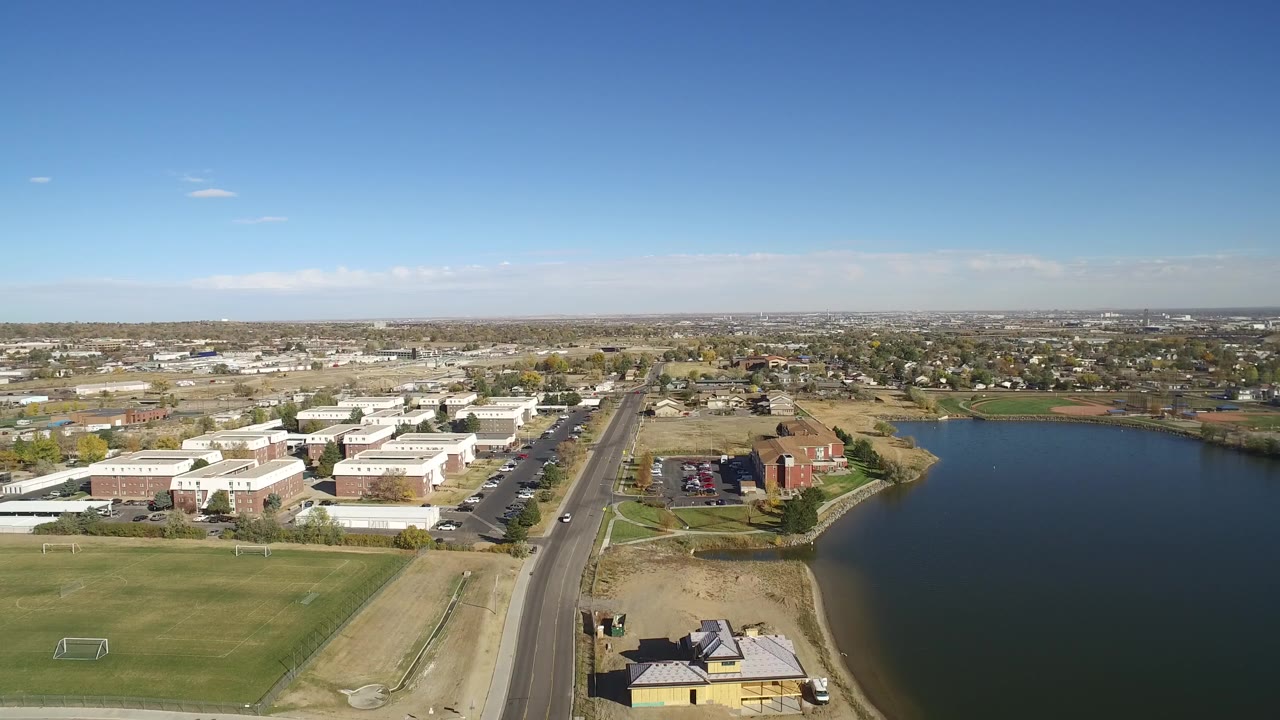 Aerial view over Hidden Lake, Colorado with a road through the center, grass fields and light-roofed buildings on the left, lake water on the right, and no visible people.