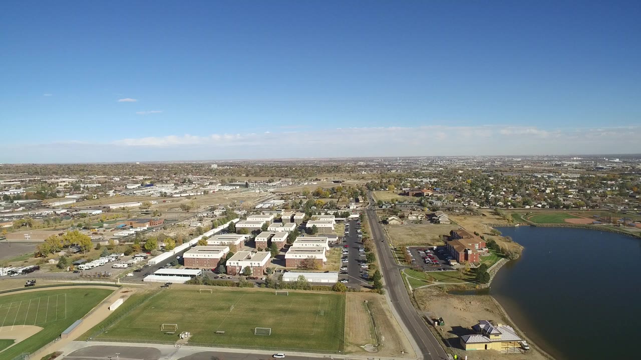 Opening aerial frame over Hidden Lake, Colorado shows green playing fields in the foreground, low-rise buildings beyond them, a road and lake at right, and no visible people.
