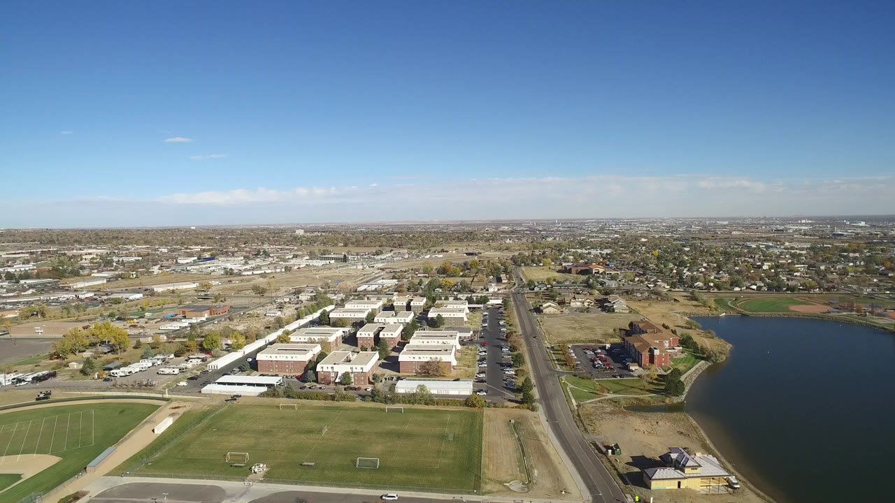 At 0:01 the Hidden Lake aerial remains wide, with the sports fields lower in frame, clustered white and brick buildings near center, the lake edge on the right, and no visible people.