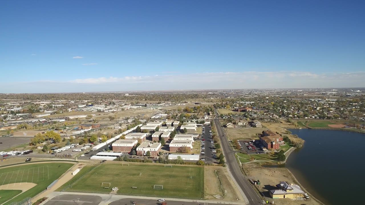 High aerial view of fields, roads, buildings, and lake with suburbs beyond.