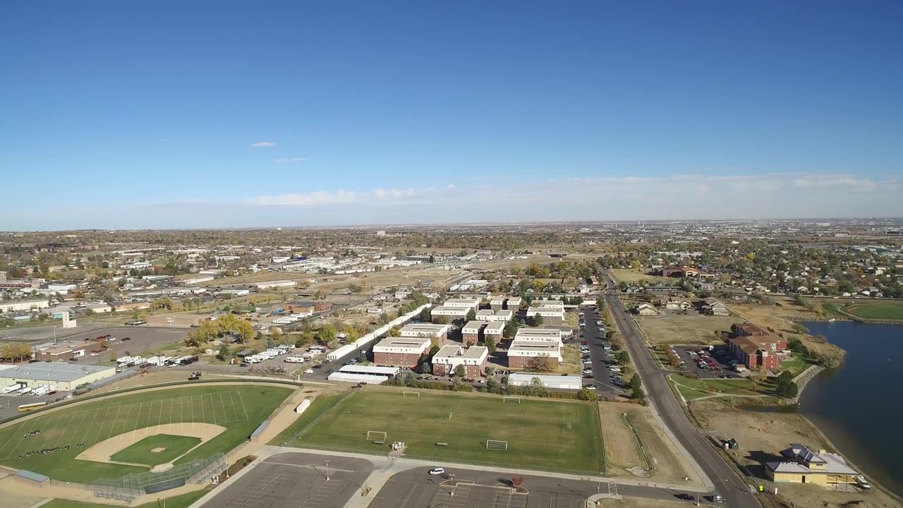 Opening aerial frame over Hidden Lake, Colorado with a baseball diamond in the foreground, low industrial buildings and homes beyond, a road along the right side, a small lake at right, and no visible people.