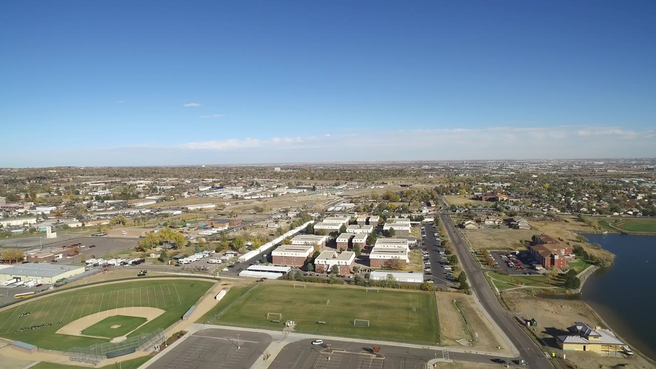 Aerial frame in Hidden Lake, Colorado showing the baseball field and empty parking lot lower in frame, warehouse-like buildings and homes across the middle, the lake edge on the right, and no visible people.