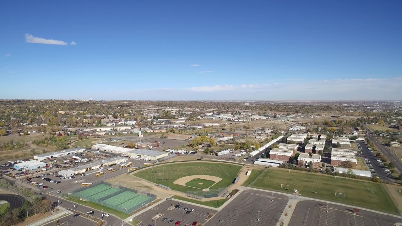 High aerial view over sports fields, parking lots, roads, and low buildings under blue sky.