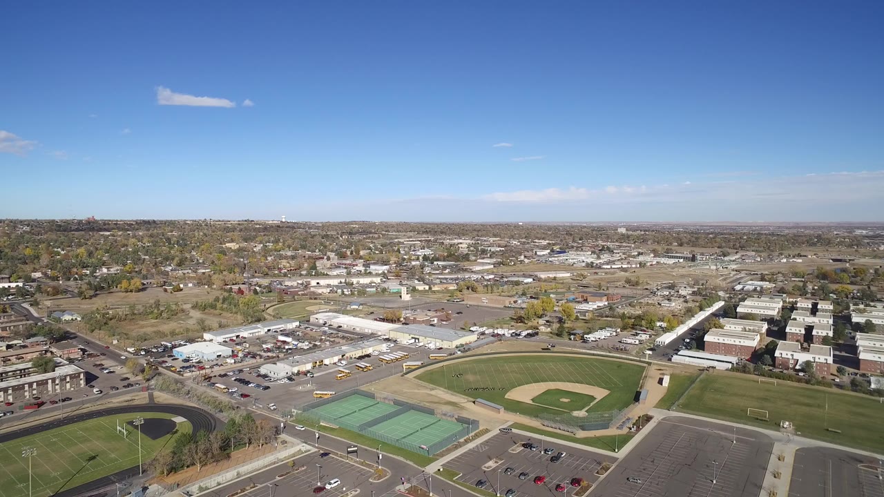 Opening aerial frame over Hidden Lake, Colorado, showing a baseball diamond, green courts, parking areas, roads, low buildings, distant neighborhoods, and no visible people.