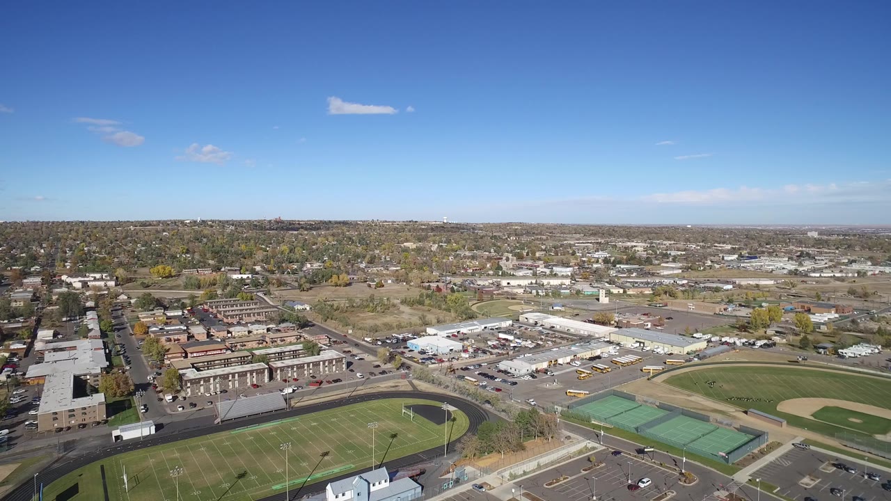 Aerial view over sports fields, tennis courts, roads, homes, and low buildings under a blue sky.