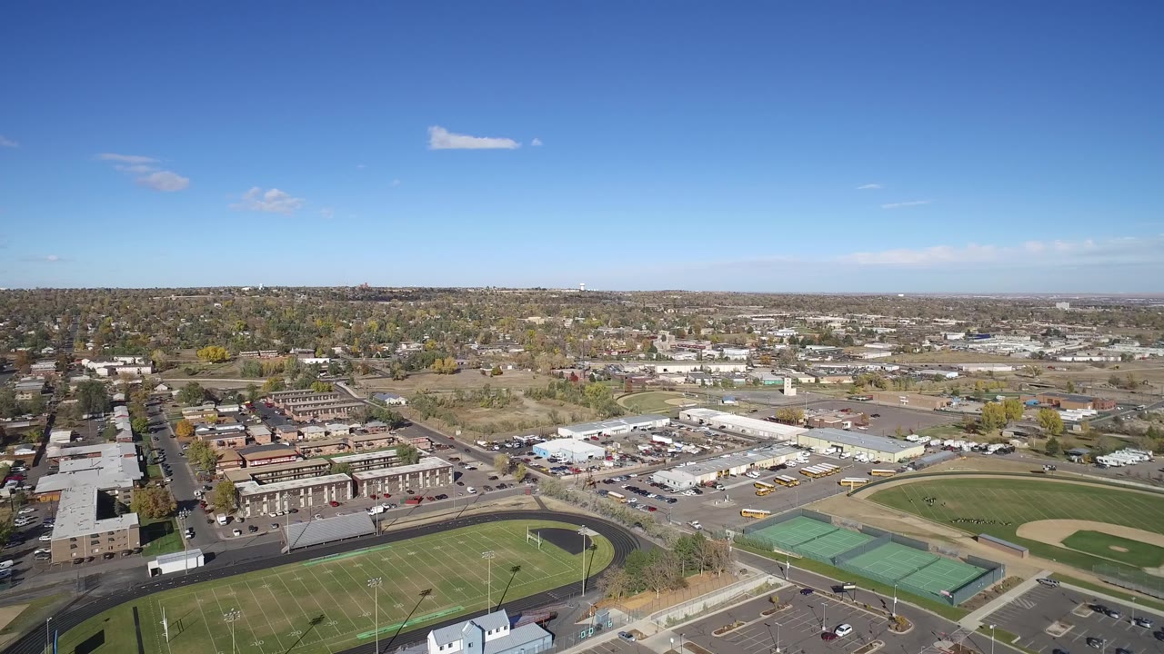 Second aerial frame in Hidden Lake, Colorado, showing the track and football field at lower left, green tennis courts near lower right, low commercial buildings in the middle distance, and no visible people.