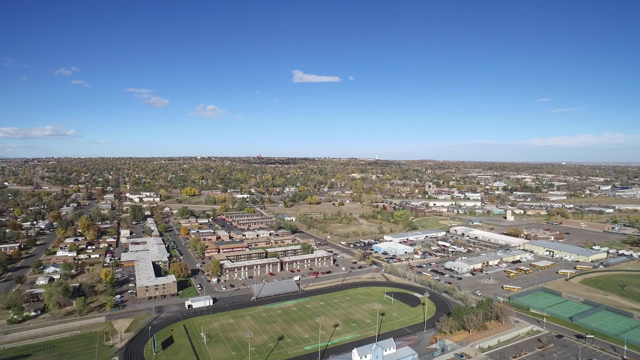 Aerial view over a school field, streets, homes, and low buildings under blue sky