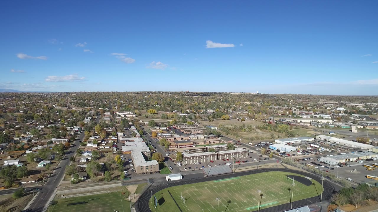 Aerial frame from Hidden Lake, Colorado with the football field and oval track in the foreground, school or commercial buildings in the midground, suburban homes beyond, and no visible people.
