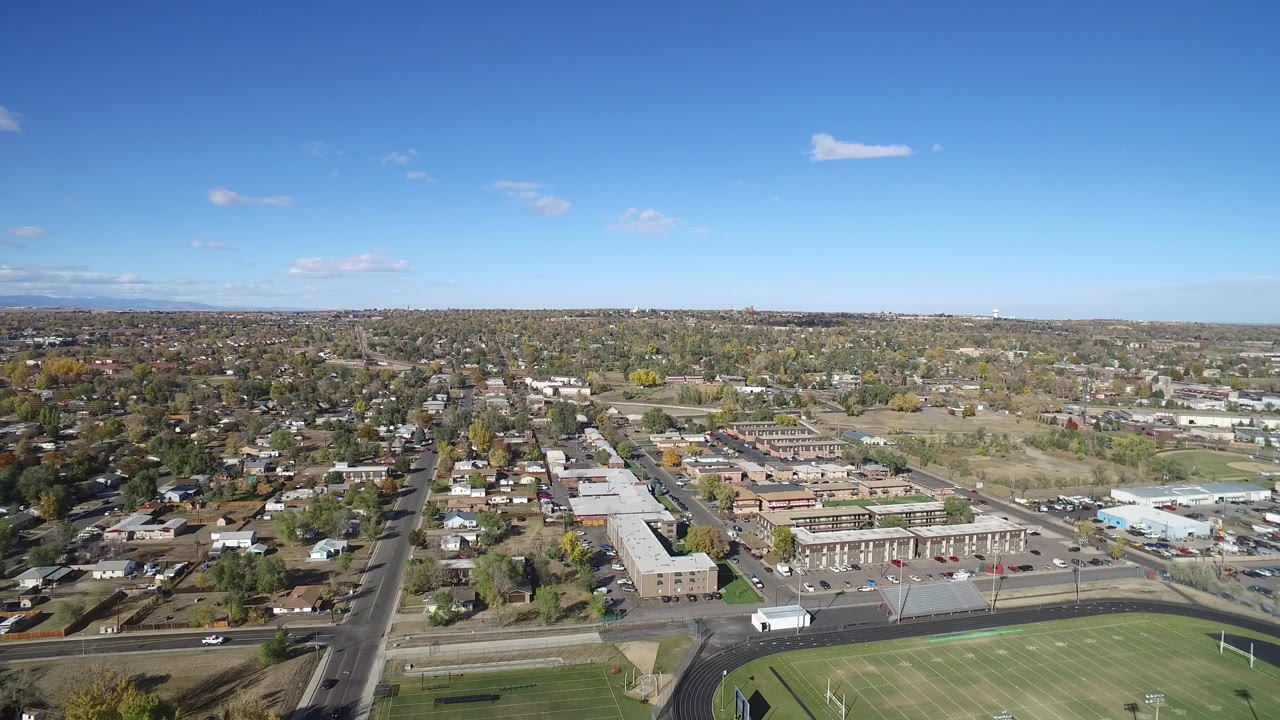 Aerial view over suburban streets, buildings, and a green sports field under blue sky.