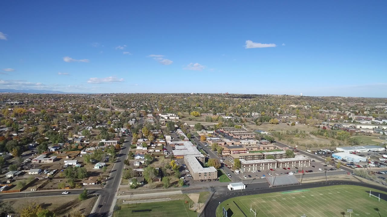 Early aerial frame of Hidden Lake, Colorado showing a central road corridor, surrounding houses, low commercial roofs, distant neighborhoods, and no visible people.