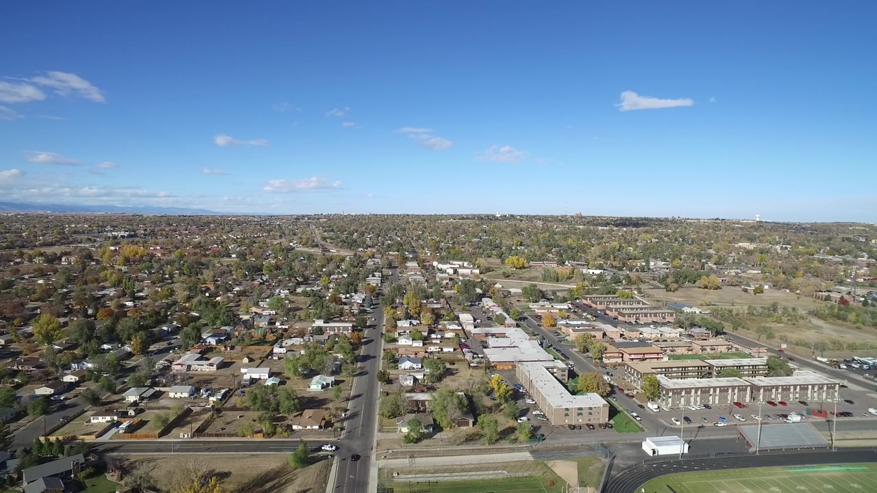 Aerial view in Hidden Lake, Colorado showing the central road more prominently, with residential roofs to the left, low buildings and parking areas to the right, and no visible people.