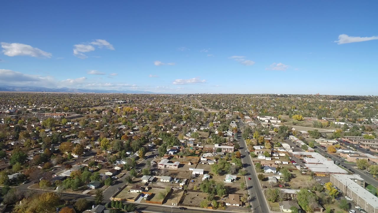 Aerial view over a residential neighborhood with roads, rooftops, trees, blue sky, and distant mountains.