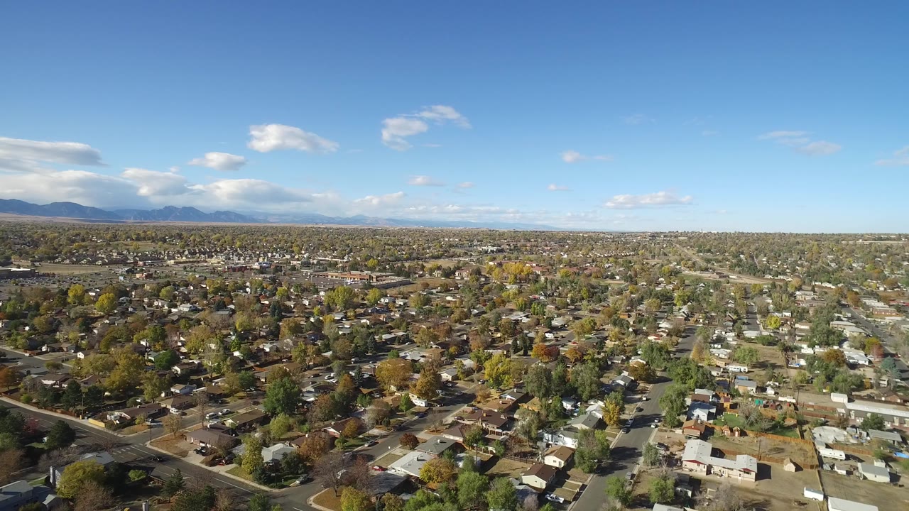 Opening frame over Hidden Lake, Colorado shows a broad residential grid, tree-lined blocks, distant mountains, scattered clouds, and no visible people.