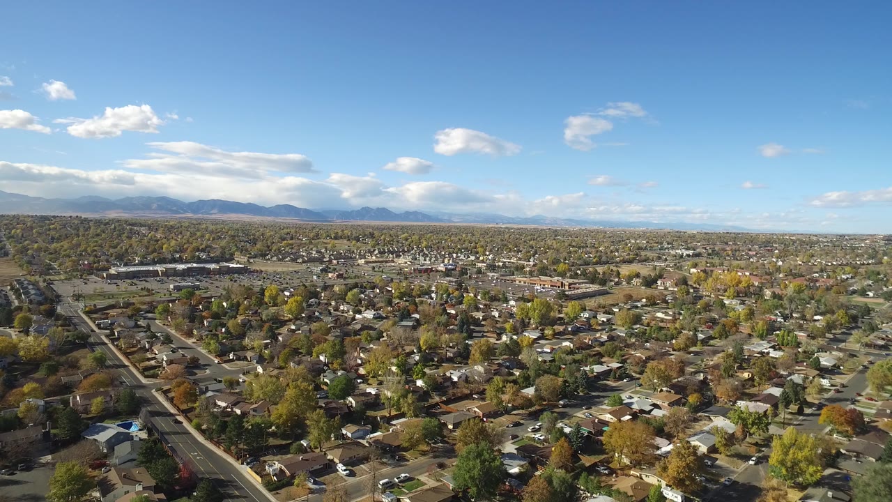 Aerial view over suburban homes, streets, trees, and distant mountains under a blue sky.