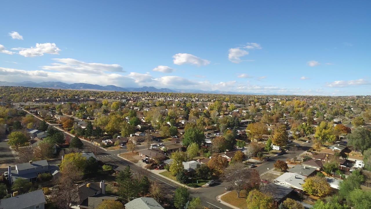 Second aerial frame near Hidden Lake, Colorado, showing a wider spread of rooftops, intersecting streets, autumn tree cover, distant mountains, and no visible people.
