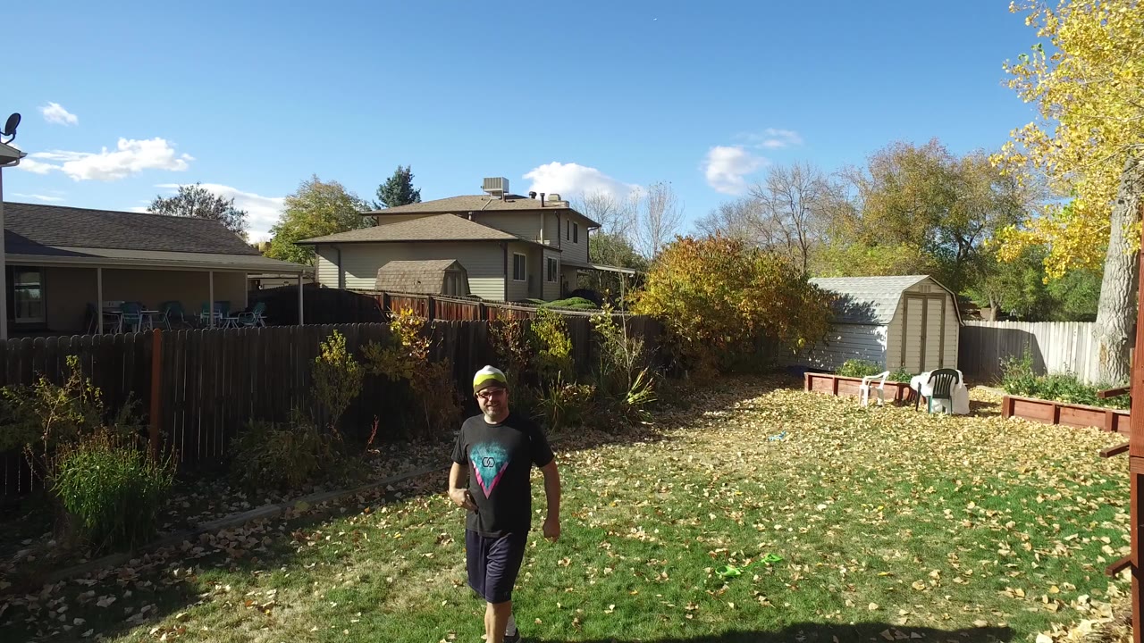 A man in a dark T-shirt and bright hat walks across a grassy backyard in Hidden Lake, Colorado, with fences, trees, a shed, and neighboring homes in the background.