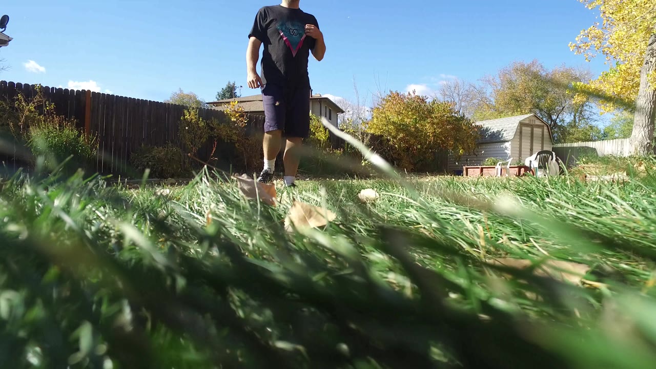 At 0:00 in Hidden Lake, Colorado, a man in a dark shirt and shorts walks toward the camera across a grassy backyard with a fence, shed, trees, and blue sky behind him.