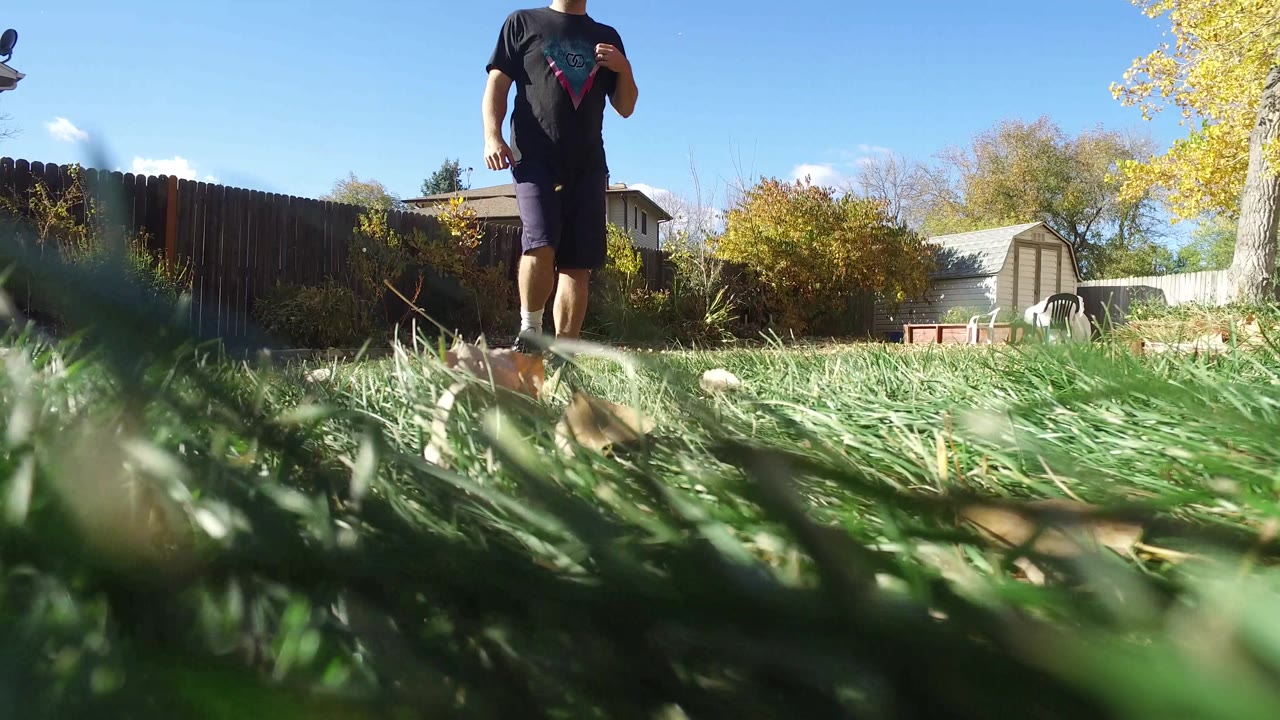 At 0:01 in Hidden Lake, Colorado, the man is closer to the low camera while tall grass fills the foreground and a wooden fence, shed, trees, and rooftops remain in the background.