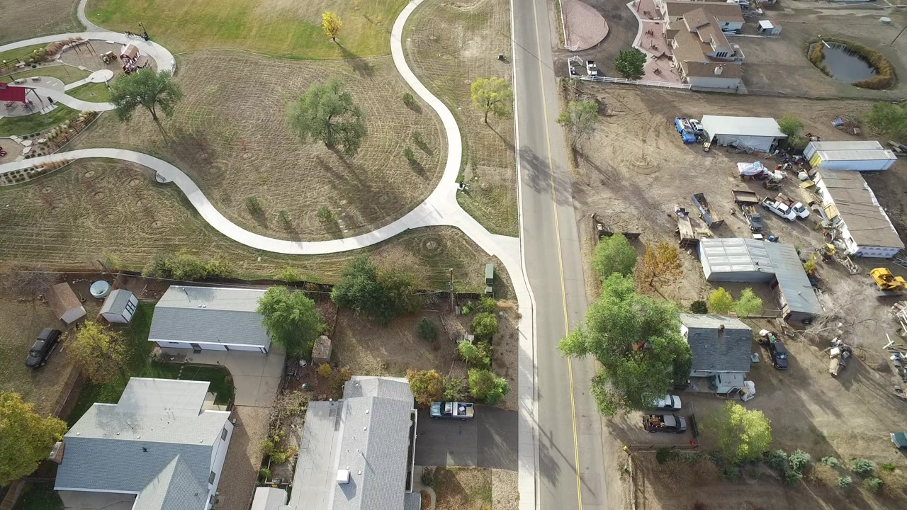 Overhead frame from Hidden Lake, Colorado showing a vertical road beside houses, trees, small sheds, and a curved light-colored path through open grass, with no visible people.