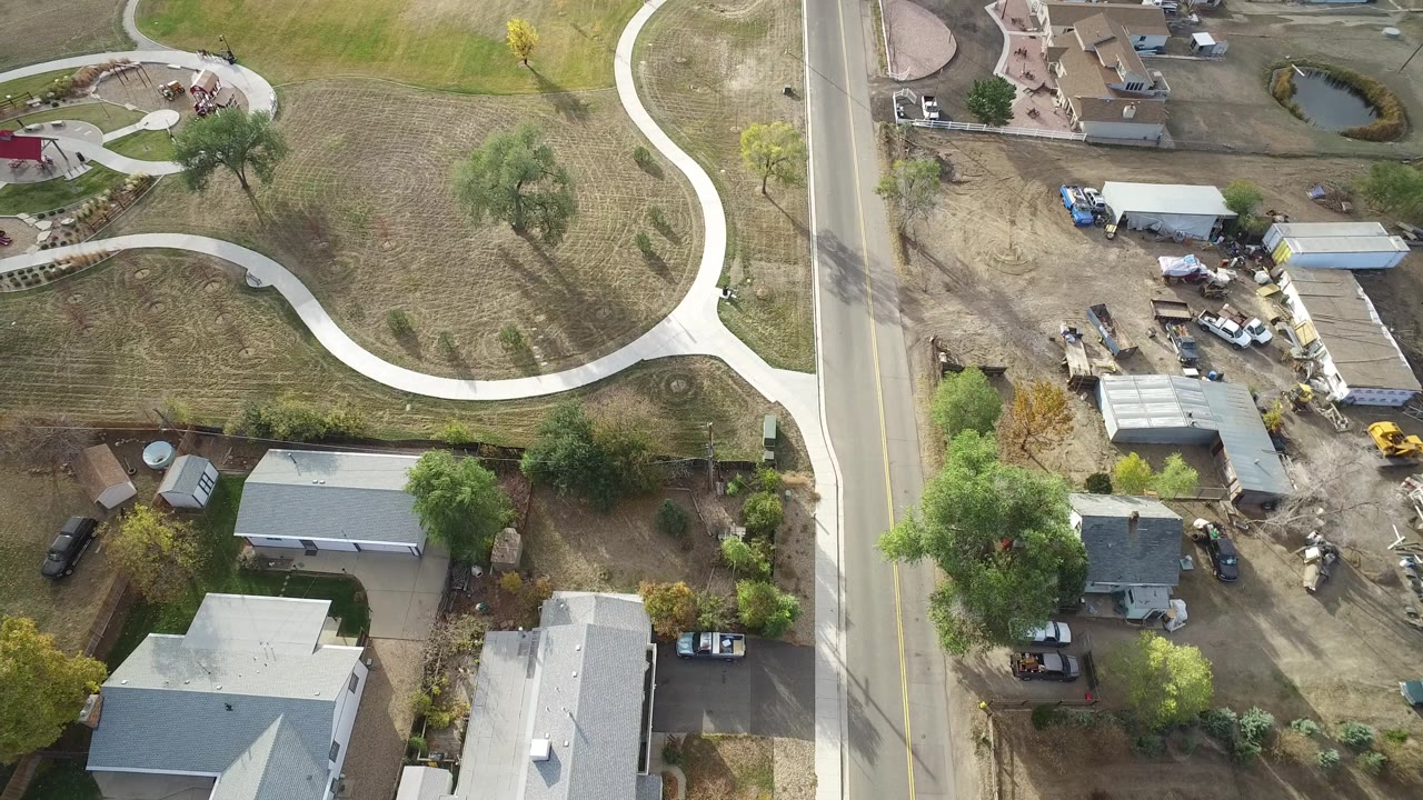 High aerial view in Hidden Lake, Colorado with the road near center, houses and work vehicles on one side, and mowed grass with a looping walkway on the other, with no visible people.