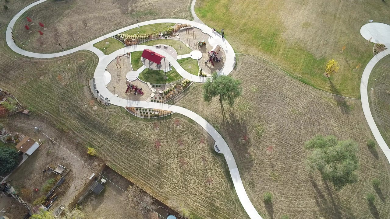 Overhead frame from Hidden Lake, Colorado showing curving white walkways around a red-roofed shelter, mown grass, small trees, garden beds, and no visible people.